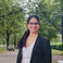 Portrait of a smiling woman with glasses and a black blazer, outdoors with green trees.