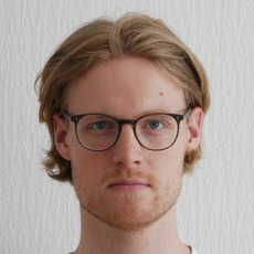 Blonde person with medium-length hair standing against a white textured wall background.
